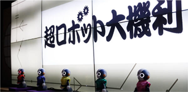 5台並んだ小さなロボット（ソータ）。後ろの画面に超ロボット大喜利と出ている写真。